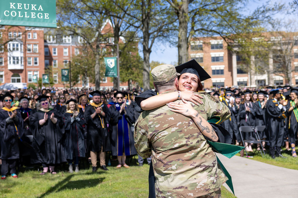 Graduate Madisyn Robinson wearing cap and gown hugging brother Tyler Robinson wearing military uniform in front of standing and clapping crowd at Commencement